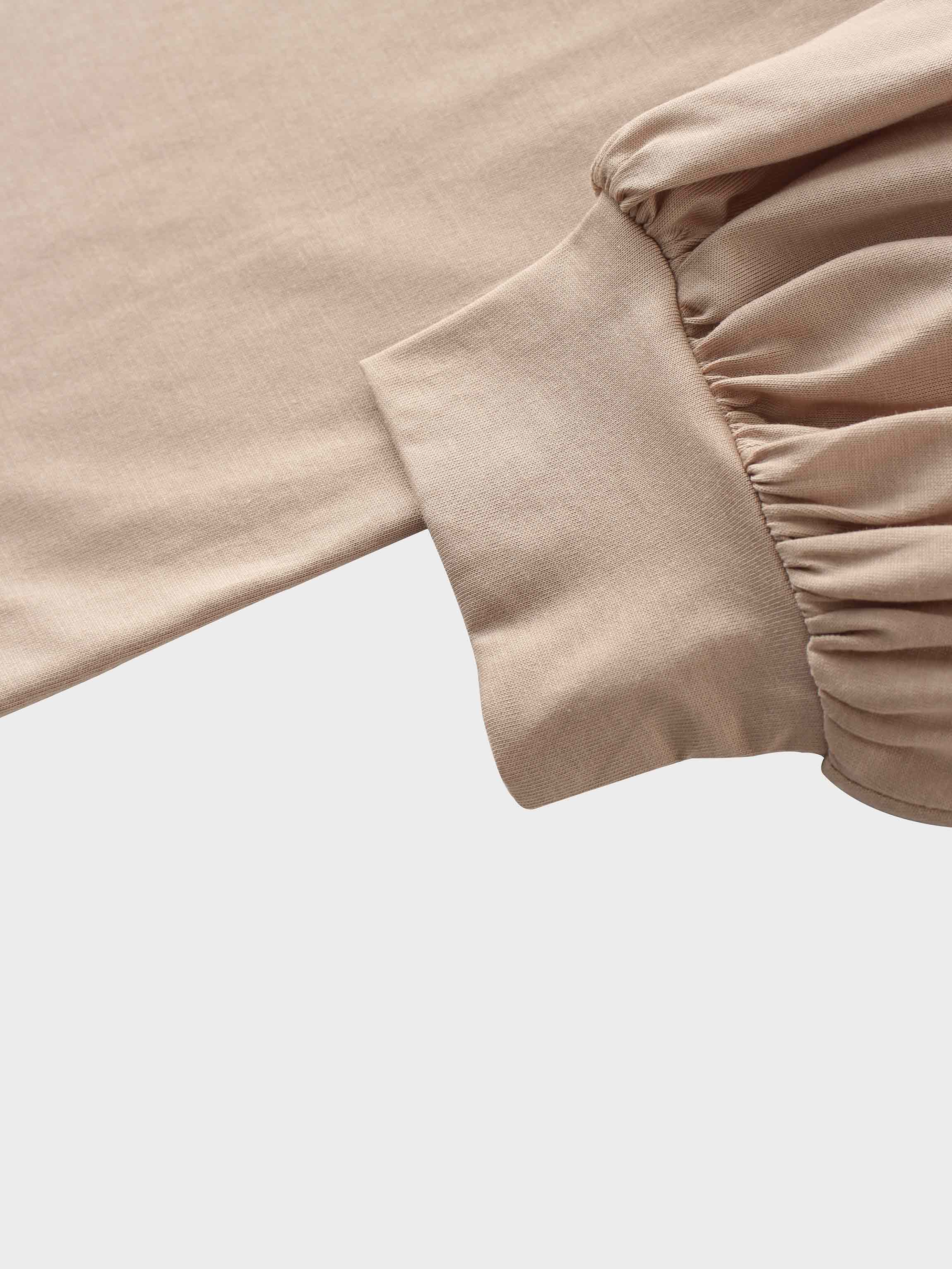 A close-up of the sleeve of the light-beige garment. The smooth fabric is gathered where it meets a wide, fitted cuff at the wrist.
